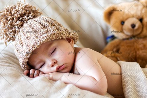 عکس با کیفیت بچه ناز خوابیده با کلاه بافتنی در کنار عروسک خرس