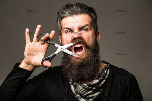 عکس مرد جوان با ریش و قیچی آرایشگری در دست بر روی صورت در پس زمینه خاکستری