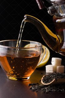 عکس ریختن چای از قوری به فنجان در کنار چای خشک در قاشق چوبی و قند و لیمو در پس زمینه مشکی