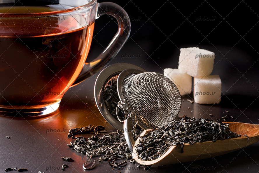 عکس صافی چای و قاشق چوبی با چای خشک در کنار فنجان چای و قند در پس زمینه مشکی