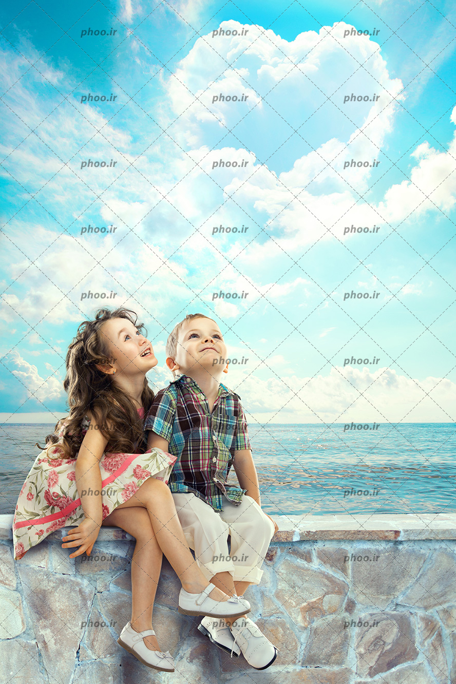 عکس با کیفیت کودکان شاد نشسته کنار دریا در حال نگاه کردن به ابر قلبی شکل در آسمان آبی