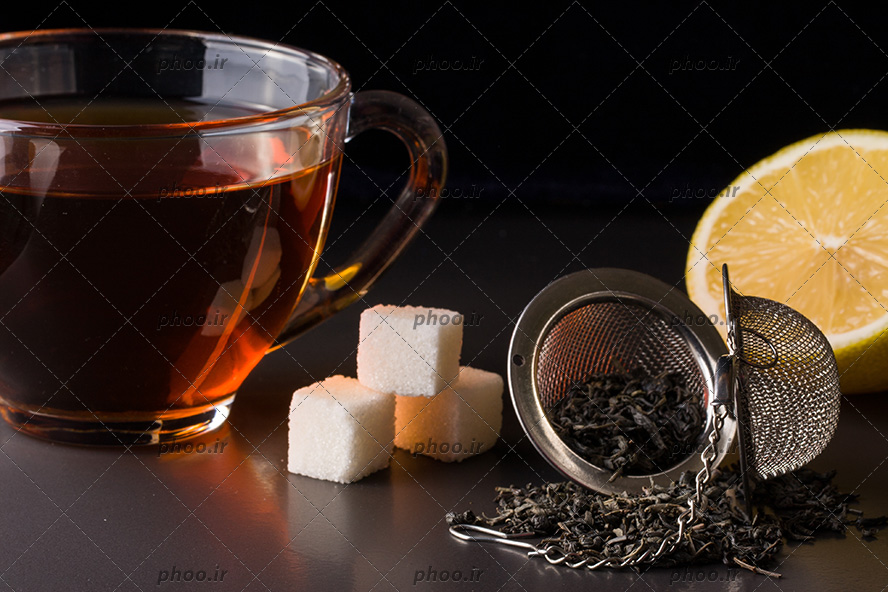 عکس چای خشک در صافی چای در کنار لیمو و قند و فنجان چای در پس زمینه مشکی