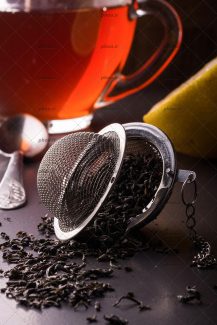 عکس چای خشک در صافی چای در کنار لیمو و فنجان چای در پس زمینه مشکی