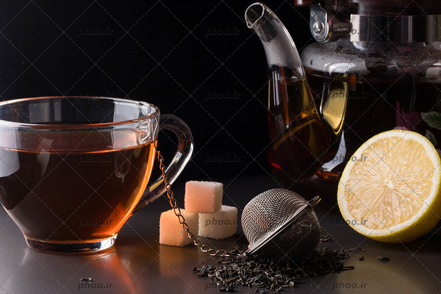 عکس چای خشک در صافی چای در کنار لیمو ، قند ، قوری و فنجان چای در پس زمینه مشکی