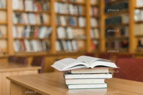 عکس با کیفیت کتاب های چیده شده بر روی یکدیگر و روی میز چوبی و قفسه کتاب ها در پس زمینه