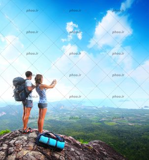 عکس با کیفیت زن و مرد گردشگر ایستاده بر روی کوه و در حال عکاسی از منظره