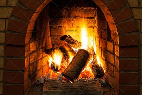 عکس چوب های در حال سوختن در شعله های آتش در شومینه ای با نمای آجری
