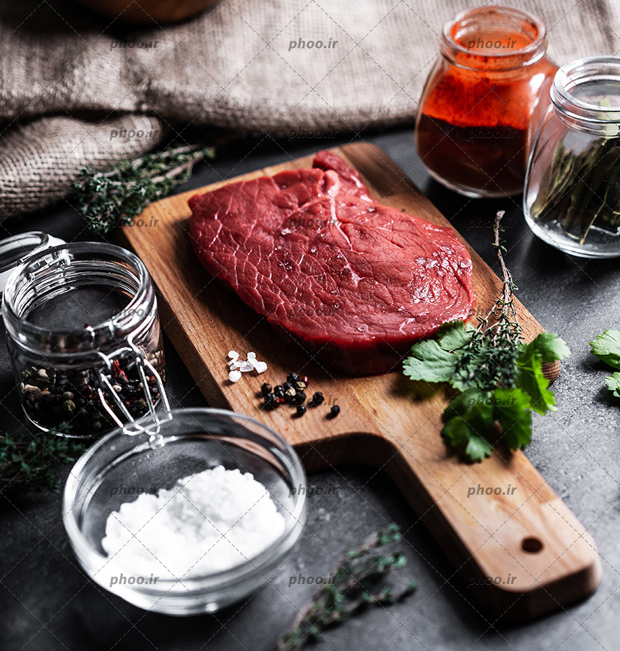 عکس گوشت روی تخته چوبی و مواد اولیه اطراف آن