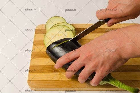 عکس دست ها در حال برش زدن بادمجان با چاقو روی تخته کار چوبی