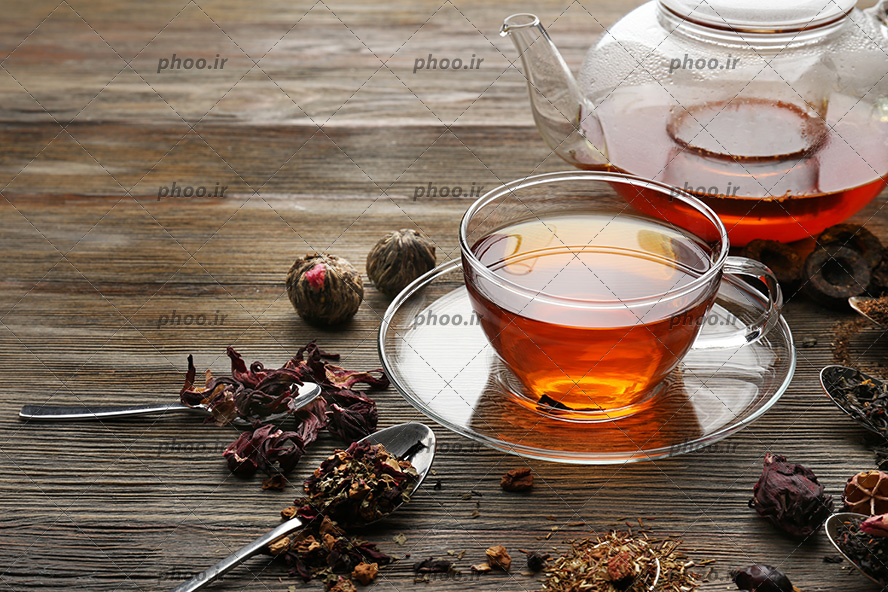 عکس چای گیاهی در قوری و فنجان و گیاه های خشک در اطراف فنجان روی زمین