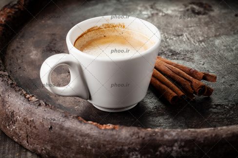 عکس فنجان سفید از قهوه در کنار چوب دارچین در سینی مشکی