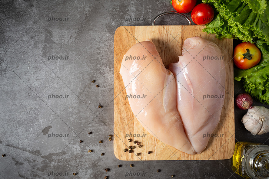 عکس با کیفیت سینه مرغ بر روی تخته چوب و سبزیجات و گوجه و سیر در کنارش