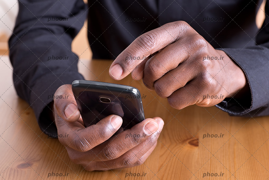 عکس مرد سیاه پوست پشت میز چوبی در حال کار با گوشی