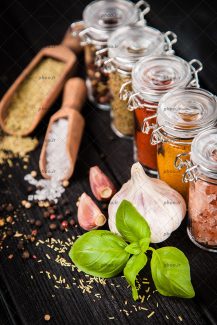 عکس ادویه هایی در ظرف های شیشه ای و روی زمین از نمای نزدیک در پس زمینه چوب مشکی