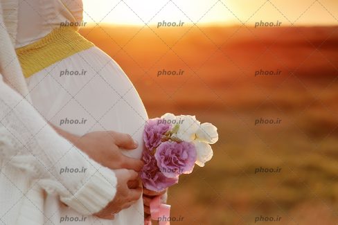 عکس با کیفیت زن باردار با یک دسته گل در برابر غروب خورشید