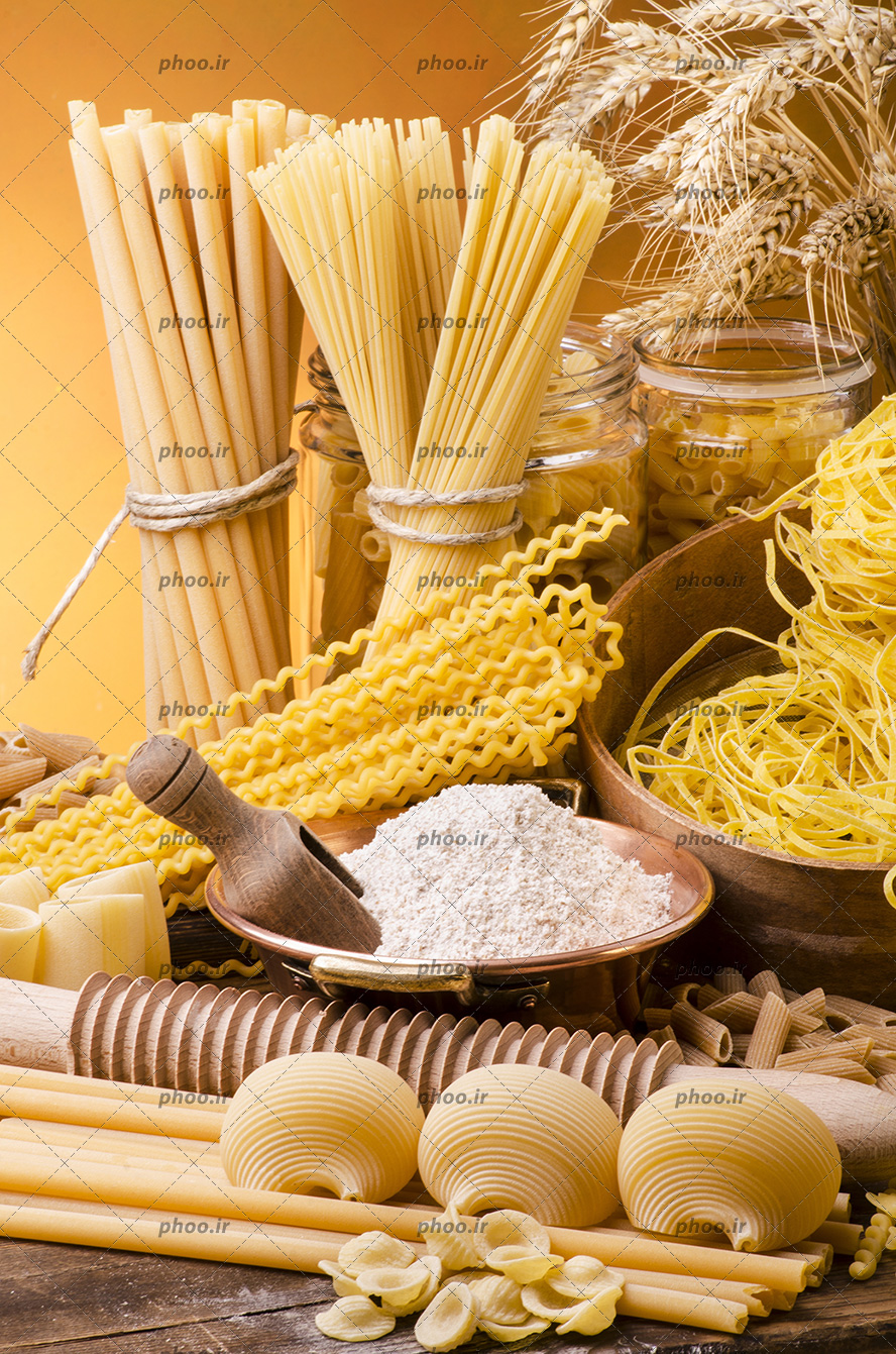 عکس ماکارونی های خام با شکل های مختلف در کنار ظرف آرد روی میز چوبی در پس زمینه قهوه ای