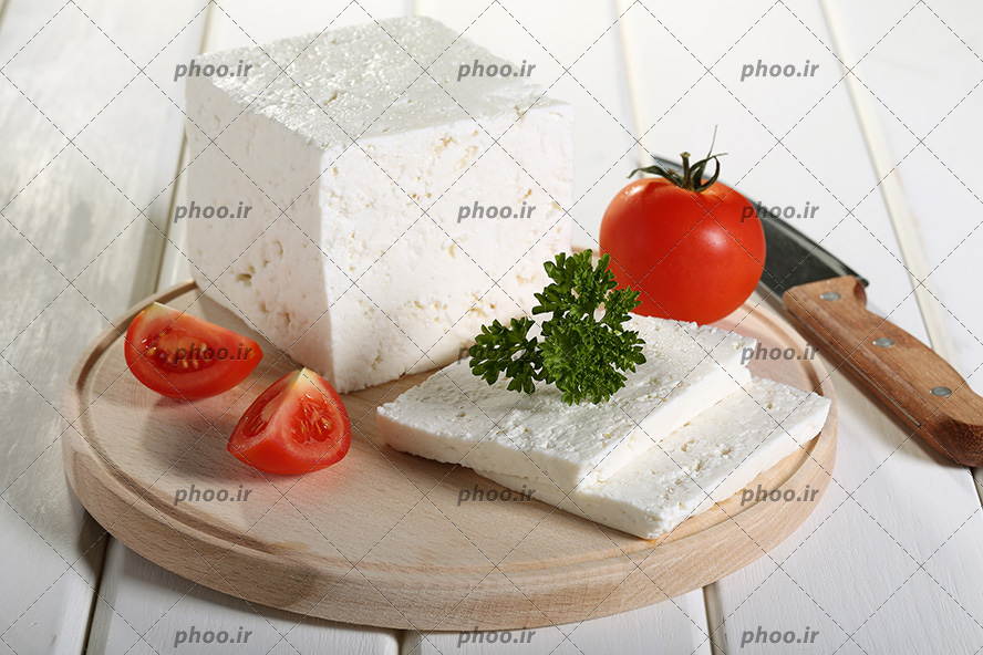 عکس یک قالب پنیر سفید و تیکه های برش خورده از آن در کنار گوجه فرنگی و سبزی روی تخته چوبی روی میز