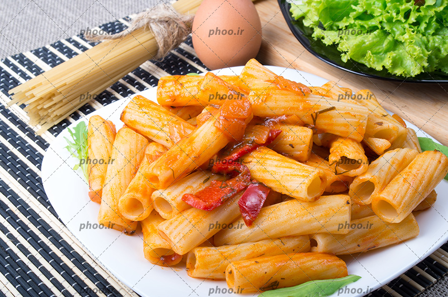 عکس ماکارونی فرمی پخته شده در بشقاب سفید در کنار تخم مرغ و ماکارونی رشته ای خام