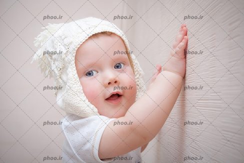 عکس نوزاد و کودک کلاه دار ایستاده با دستهایش کنار دیوار