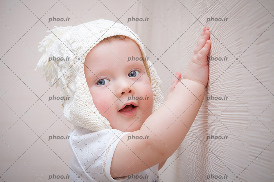 عکس نوزاد و کودک کلاه دار ایستاده با دستهایش کنار دیوار