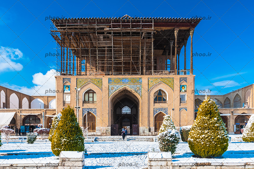 عکس یک مکان قدیمی که با آجر ساخته شده و کاشی کاری زیبا و در و پنجره های چوبی و همچنین عکس امام خمینی (ره) و امام خامنه ای در بالای ساختمان گذاشته شده و طبقه دوم در حال ساختن است و درختانی که برف روی آن نشسته
