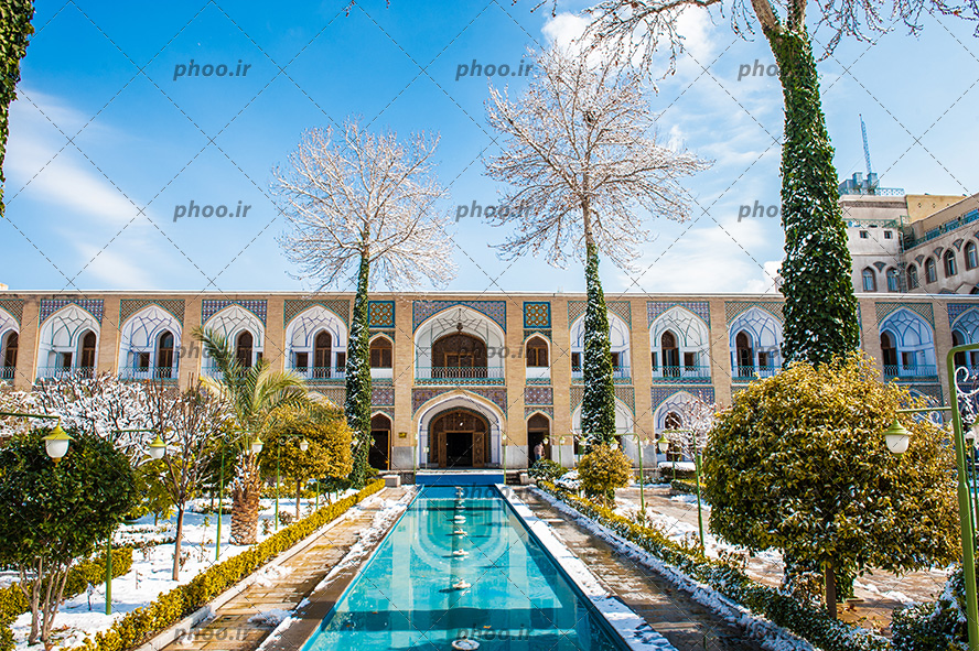 عکس یک خانه قدیمی با بالکن های زیاد و در و پنجره های چوبی و در حیاط آن حوض آب و درختانی که روی آن برف نشسته دارد