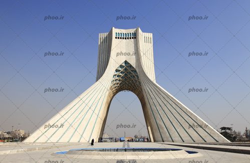 عکس برج آزادی نمای سنگ شده و آسمانی آبی و شهری که در پشت آن دیده می شود