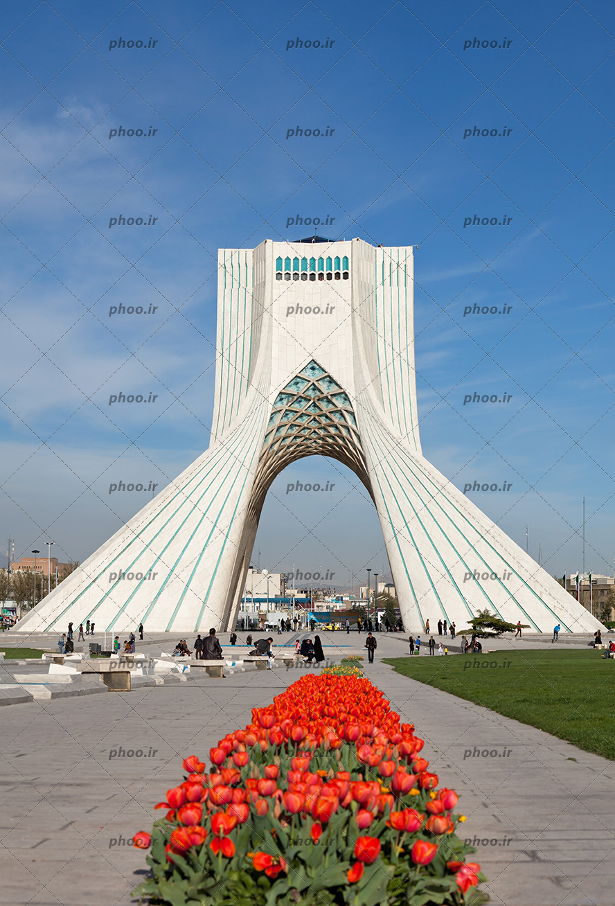 عکس برج آزادی نمای سنگ شده و آسمانی آبی و شهری که در پشت آن دیده می شود و بازدید کننده هایی که به آنجا آمدند و گل های لاله قرمز
