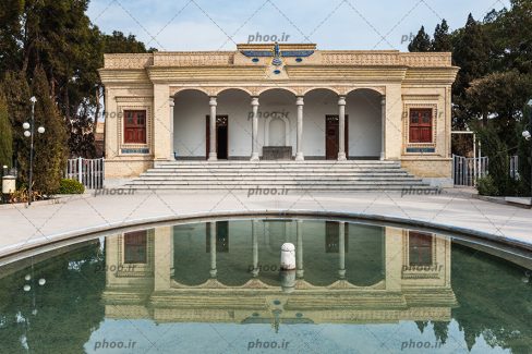 عکس یک خانه با معماری زیبا و ستون های جلوی آن و در و پنجره های چوبی مستطیلی آن و حوضی که در حیاط آن است و انعکاس عکس خانه در دل آب حوض