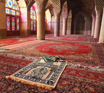 عکس از داخل مسجد با دیوار ها و ستون هایی که مرمت کاری شده است و فرش های قرمز کف و پنجره هایی با شیشه های رنگی و سجاده در انتظار نماز