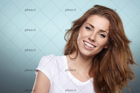 عکس با کیفیت زن با لبخند و مو های قهوه ای و تی شرت سفید در پس زمینه آبی