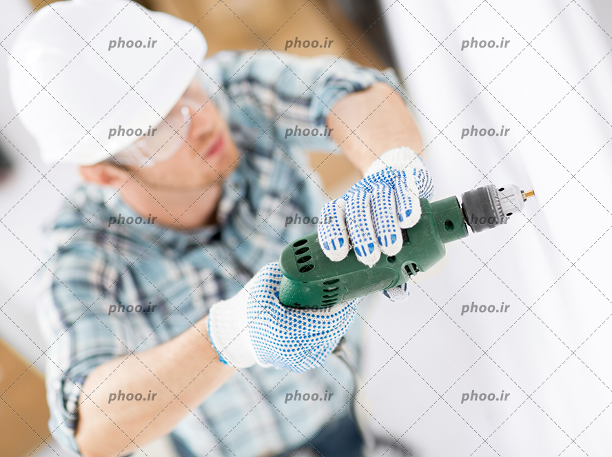 عکس کارگر با کلاه ایمنی و دستکش و مته برقی در حال سوراخ کردن دیوار