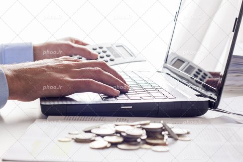 عکس با کیفیت مرد در حال کار کردن با لپ تاپ و ماشین حساب و چند سکه طلایی در کنار لپ تاپش