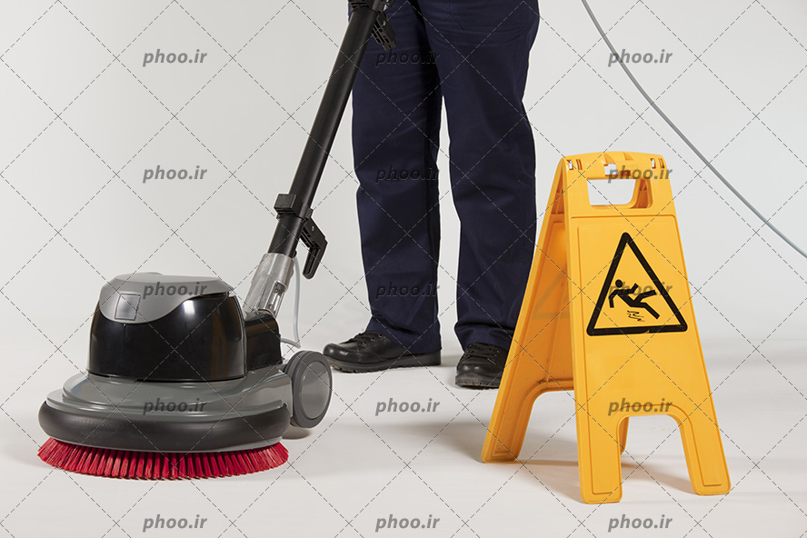 عکس کارگر در حال تمیز کردن کف با دستگاه کف شوی و یک تابلو زرد برای هشدار
