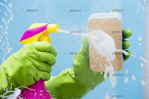 عکس با کیفیت نمایی از تمیز کردن شیشه با اسفنج و شیشه شوی توسط بانویی با دستکش پلاستیکی سبز رنگ
