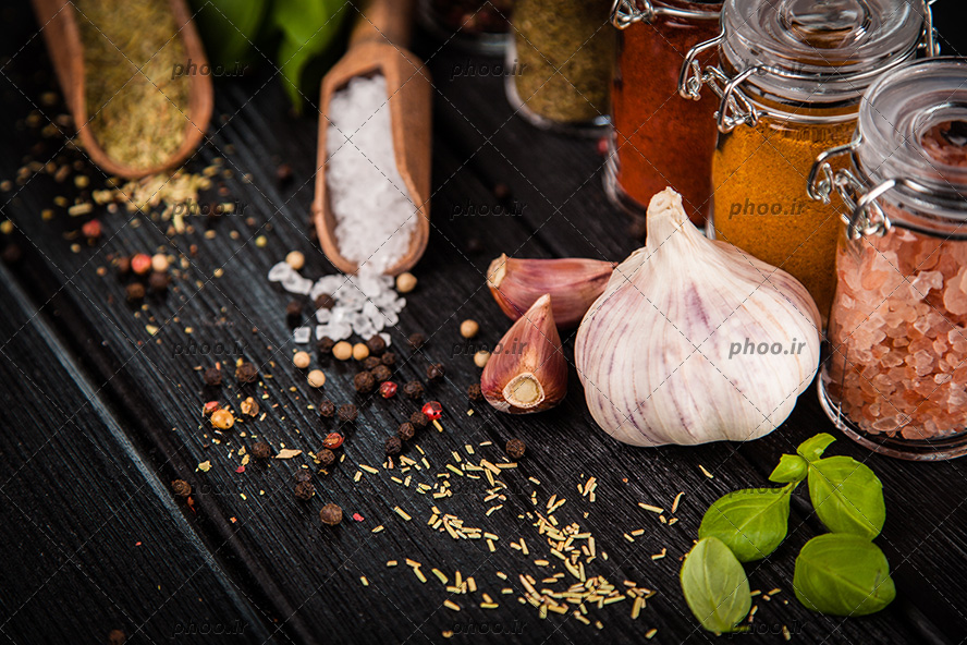 عکس ادویه های متفات روی زمین و در قاشق های چوبی و ظرف های شیشه ای در پس زمینه چوب مشکی
