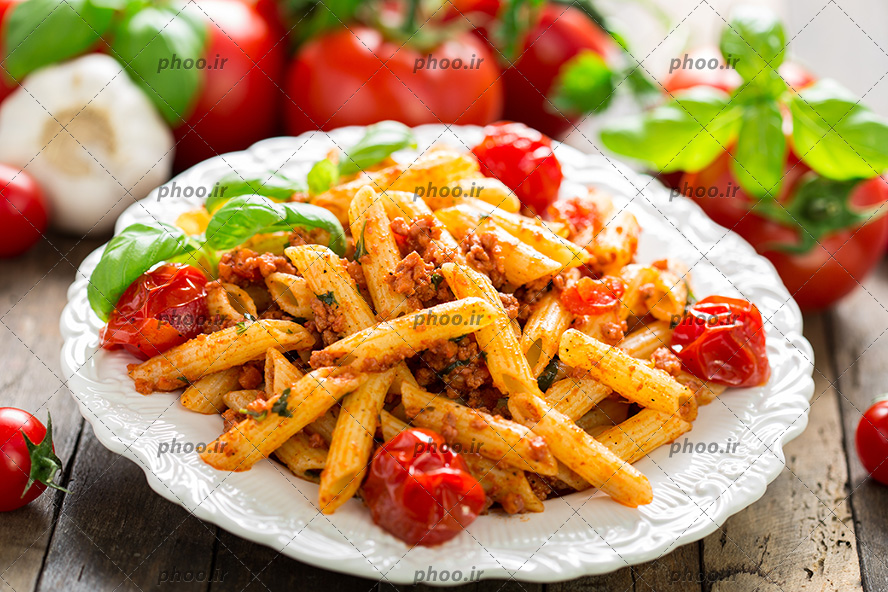 عکس پاستا با سس گوجه فرنگی و برگ ریحان تازه در بشقاب در کنار گوجه و سیر در پس زمینه چوبی