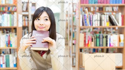 عکس دختر در کتابخانه نشسته پشت میز با یک کتاب در دست