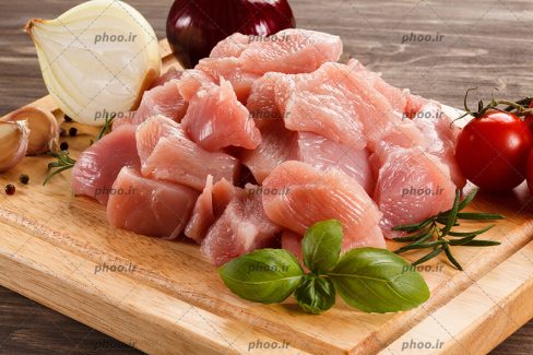 عکس مرغ تکه شده در کنار سبزی و گوجه روی تخته چوبی