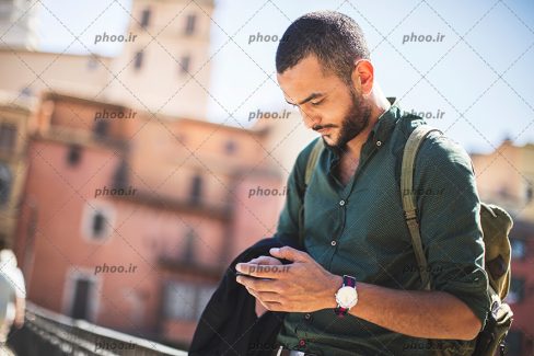 عکس مرد جوان با ریش و کوله پشتی در حال کار با گوشی در شهر