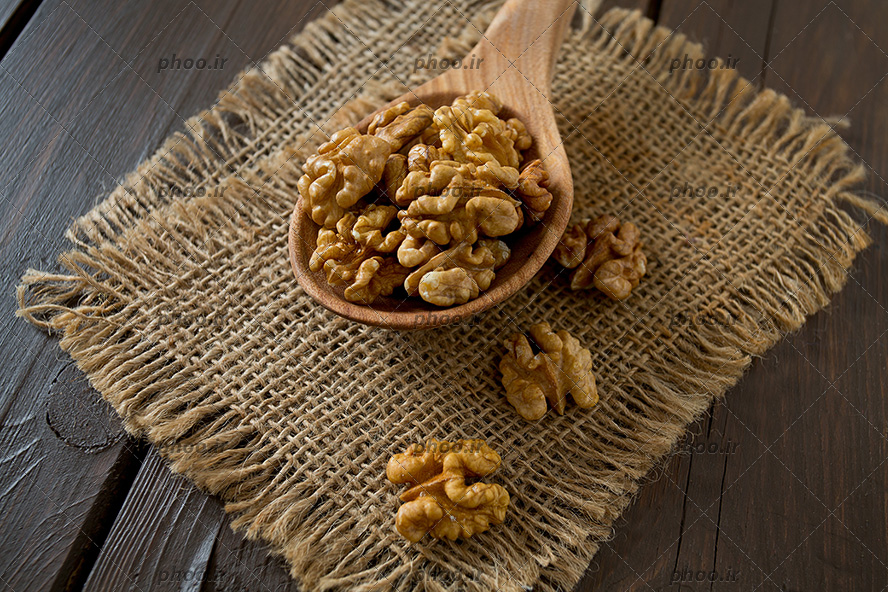 عکس مغز گردو در قاشق چوبی روی پارچه کنفی در پس زمینه چوبی