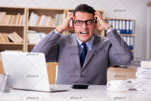 عکس مرد عصبانی در دفتر کار نشسته پشت میز و ماشین حساب و لپ تاپ جلوی او