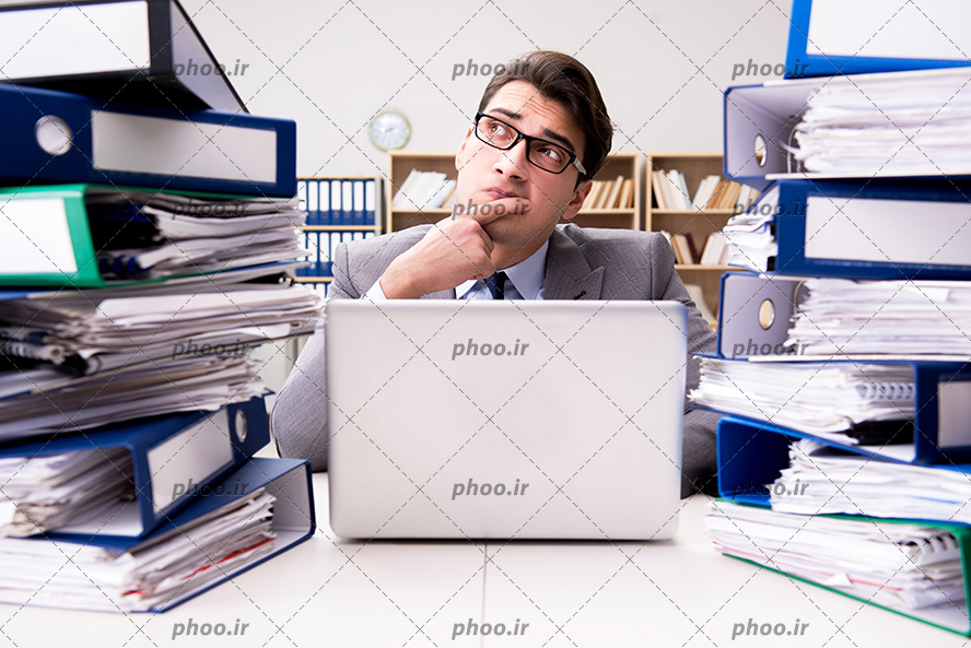 عکس تاجر در دفتر کار در حال فکر کردن در مقابل پرونده ها و لپ تاپ روی میز