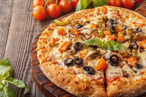 عکس پیتزا با مخلفات در کنار ریحان و گوجه در پس زمینه چوبی