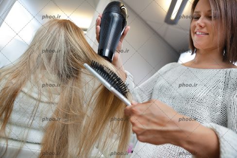 عکس آرایشگر در حال سشوار کردن مو های خانم مشتری در آرایشگاه