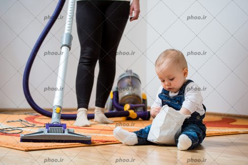 عکس مادر در حال تمیز کردن اتاق با جاروبرقی و کودک روی زمین نشسته است