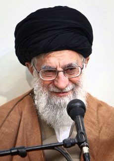 عکس با کیفیت مقام معظم رهبری با عمامه مشکی و عبا به رنگ قهوه ای در حال لبخند زدن و میکروفون در مقابل صورت ایشان