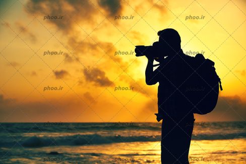 عکس با کیفیت مرد عکاس با دوربین عکاسی در کنار دریا و در مقابل غروب خورشید