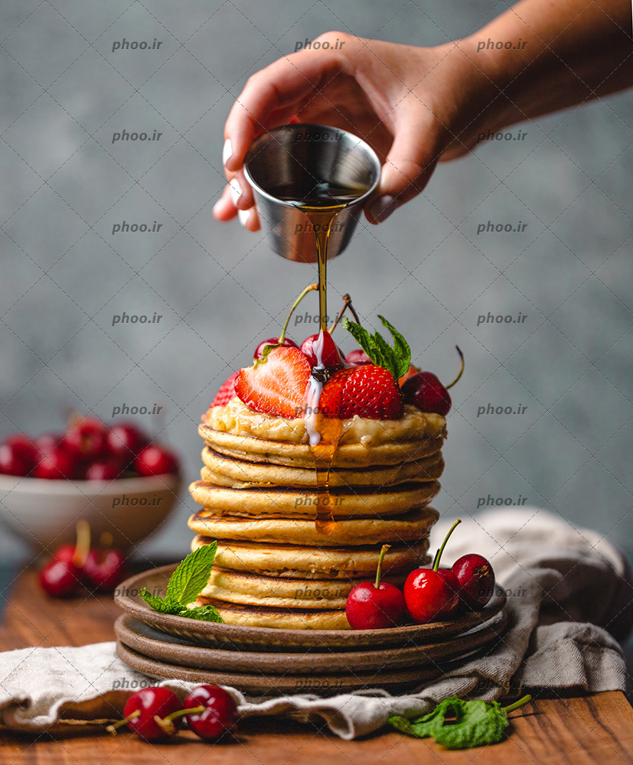 عکس شیرینی پنکیک های روی هم با عسل درحال ریختن و تاپینگ توت فرنگی و گیلاس