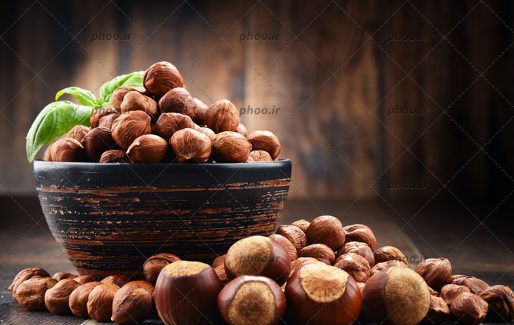 عکس فندق هایی داخل کاسه با یک برگ ریحان و روی زمین در پس زمینه چوبی